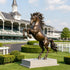 Large bronze rearing horse sculpture displayed on a stone pedestal at an elegant equestrian racetrack, featuring raised front legs, powerful muscular anatomy, a dramatically flowing mane and tail, and a warm antique bronze patina.