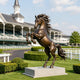 Large bronze rearing horse sculpture displayed on a stone pedestal at an elegant equestrian racetrack, featuring raised front legs, powerful muscular anatomy, a dramatically flowing mane and tail, and a warm antique bronze patina.