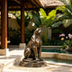 Large bronze seated panther sculpture beside a tropical resort pool, featuring a poised upright posture, muscular body, curled tail, realistic feline face, and warm bronze patina on a natural rock-style base.