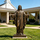 Life-size bronze Jesus statue with open arms and flowing robes displayed on a pedestal in a landscaped church garden under a clear blue sky.