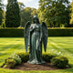 Life-size bronze guardian angel statue with large feathered wings and flowing robes, displayed among flowers and manicured shrubs in a peaceful garden setting.
