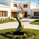 Large outdoor abstract bronze sculpture with flowing green patina and golden curves surrounding a white sphere, installed on a black pedestal in the landscaped front garden of a modern luxury villa.