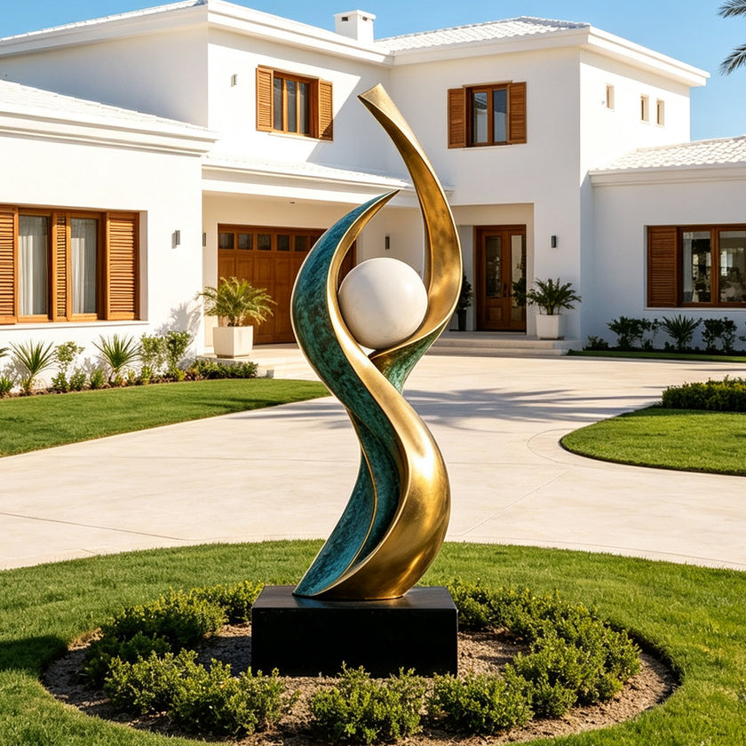 Large outdoor abstract bronze sculpture with flowing green patina and golden curves surrounding a white sphere, installed on a black pedestal in the landscaped front garden of a modern luxury villa.
