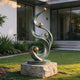 Contemporary abstract bronze garden sculpture with flowing verdigris curves and polished golden spheres, displayed on a natural stone pedestal beside a modern residence.