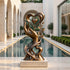 Elegant bronze hand and heart sculpture displayed on a light stone pedestal beside a reflecting pool in a luxury modern courtyard, featuring an upward-reaching hand, layered heart shapes, delicate leafy branches, and flowing ribbon forms with a warm antique bronze finish.