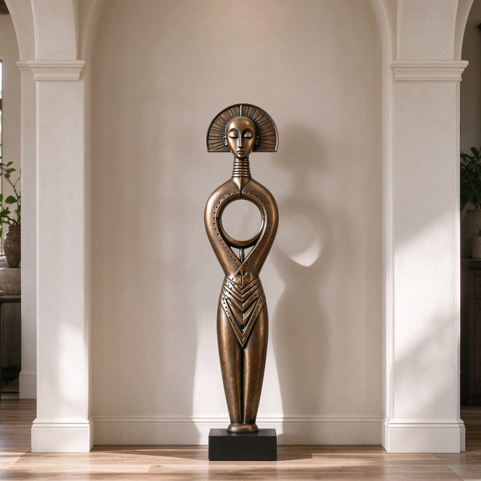 A serene bronze floor sculpture of a tribal goddess in a modern hallway. The tall figure features a sunburst halo headdress, a closed-eye meditative face, and a distinctive circular hollow torso. This antique-finished metal art piece adds cultural elegance and peaceful balance to the indoor space.