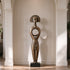 A serene bronze floor sculpture of a tribal goddess in a modern hallway. The tall figure features a sunburst halo headdress, a closed-eye meditative face, and a distinctive circular hollow torso. This antique-finished metal art piece adds cultural elegance and peaceful balance to the indoor space.