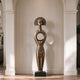 A serene bronze floor sculpture of a tribal goddess in a modern hallway. The tall figure features a sunburst halo headdress, a closed-eye meditative face, and a distinctive circular hollow torso. This antique-finished metal art piece adds cultural elegance and peaceful balance to the indoor space.