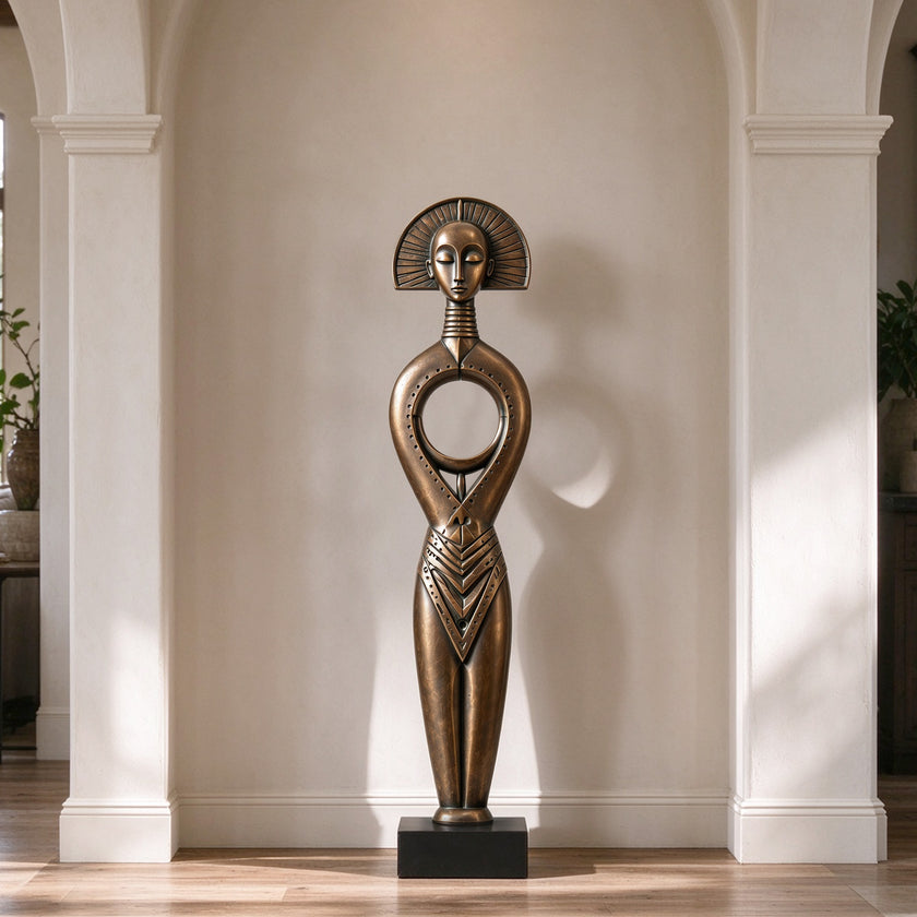 A serene bronze floor sculpture of a tribal goddess in a modern hallway. The tall figure features a sunburst halo headdress, a closed-eye meditative face, and a distinctive circular hollow torso. This antique-finished metal art piece adds cultural elegance and peaceful balance to the indoor space.