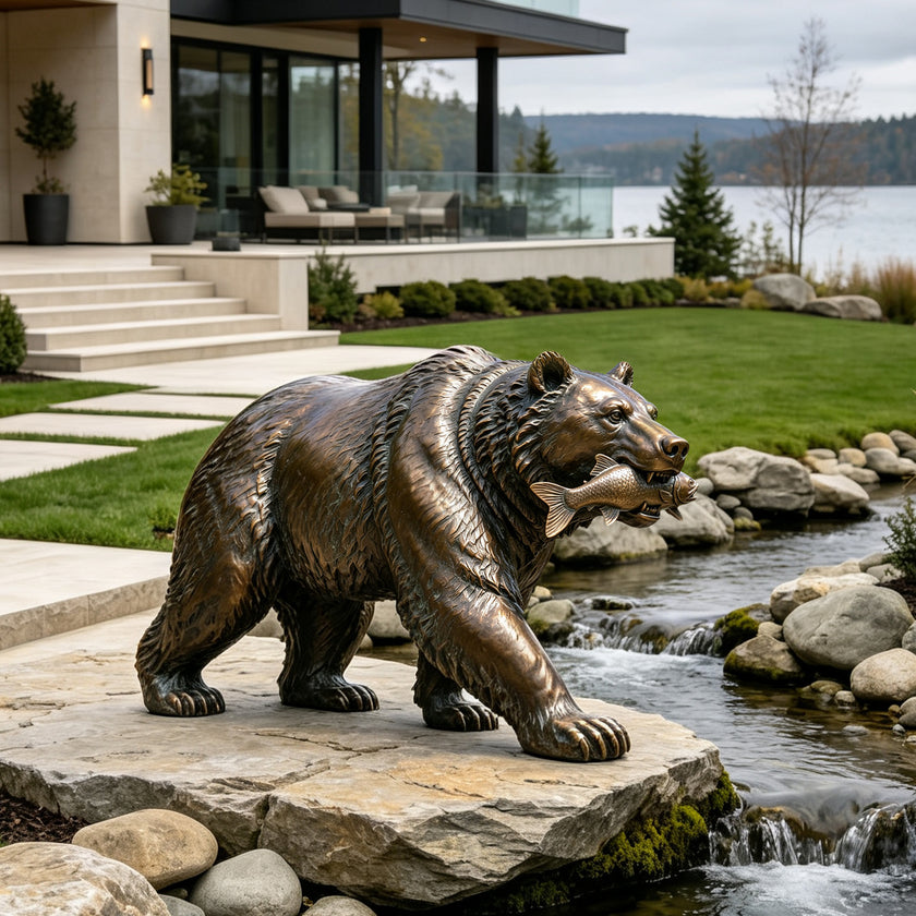 A realistic cast bronze sculpture of a grizzly bear holding a fish in its mouth. The highly textured wildlife metal art features a dynamic hunting pose, set beautifully next to a rocky backyard stream with a lakeside view.