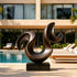 A large bronze sculpture featuring a dynamic, sweeping wave-like curve. This fluid abstract metal art piece is displayed on a sunny stone patio next to a pristine swimming pool and modern lounge chairs, adding luxury to the villa exterior.