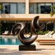 A large bronze sculpture featuring a dynamic, sweeping wave-like curve. This fluid abstract metal art piece is displayed on a sunny stone patio next to a pristine swimming pool and modern lounge chairs, adding luxury to the villa exterior.