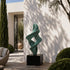 A large bronze sculpture with a green patina, featuring an abstract winding flame or twisting spiral design. This elegant modern metal art piece is displayed outdoors in a minimalist courtyard against a sunlit white wall.
