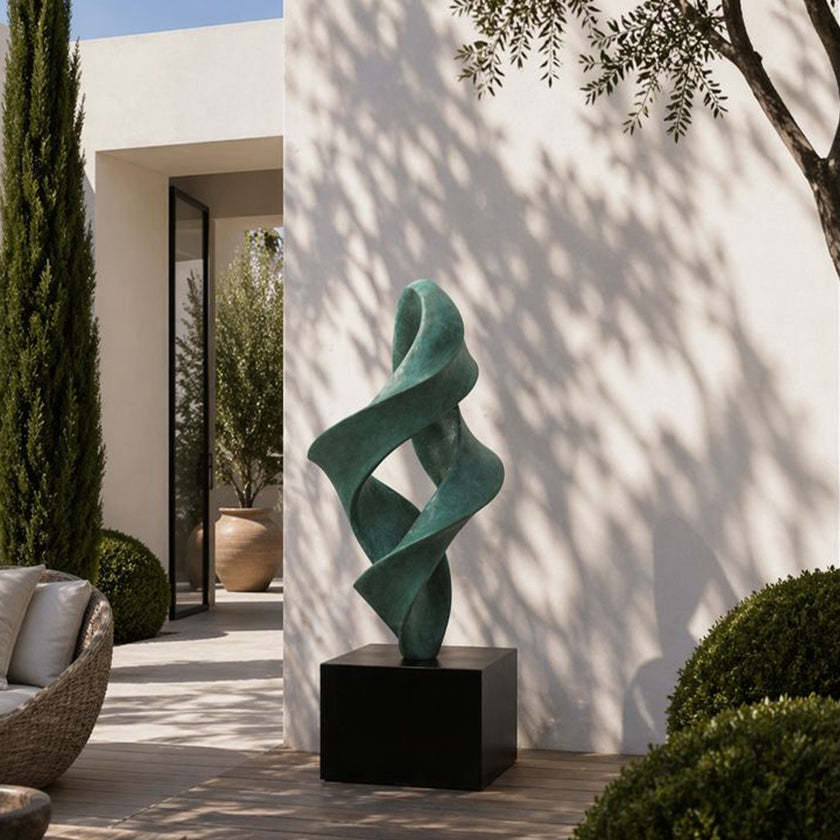 A large bronze sculpture with a green patina, featuring an abstract winding flame or twisting spiral design. This elegant modern metal art piece is displayed outdoors in a minimalist courtyard against a sunlit white wall.