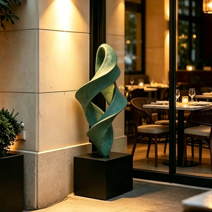 A large, abstract green bronze sculpture with a twisting, flame-like design stands on a black square pedestal. It is located outdoors next to a light stone wall, just outside the glass doors of an elegantly lit restaurant.