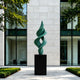 A large bronze sculpture featuring an abstract, intertwining green spire with a double helix design. This sleek, modern metal art piece is beautifully displayed outdoors in front of a symmetrical luxury office building with glass windows.