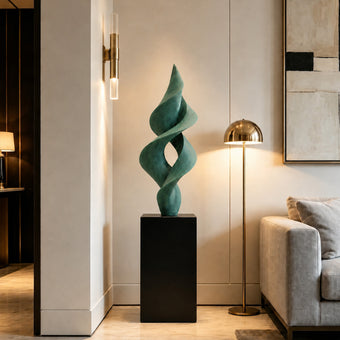 A twisting, abstract green sculpture on a black pedestal in a modern interior, placed beside a brass floor lamp and a grey sofa