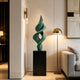 A twisting, abstract green sculpture on a black pedestal in a modern interior, placed beside a brass floor lamp and a grey sofa