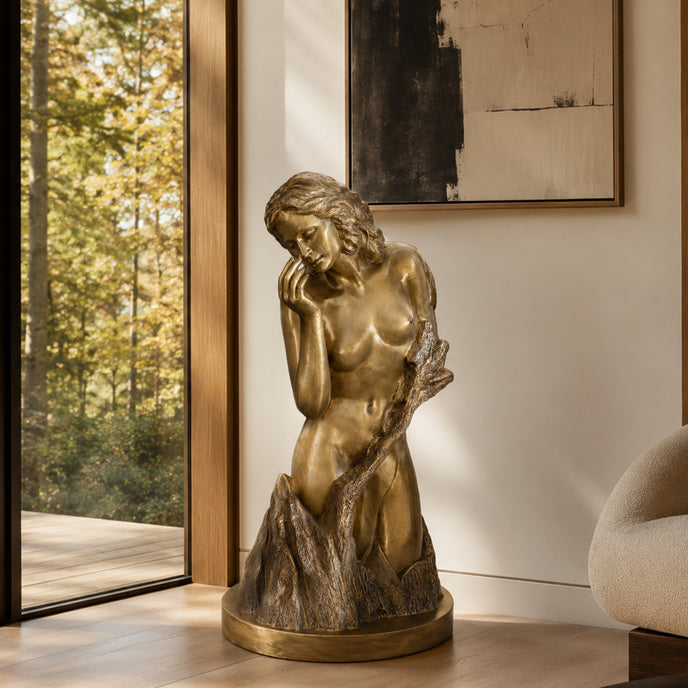 A bronze statue of a pensive female figure with wavy hair, gently touching her cheek, emerging from wave-like rock textures. This elegant metal art piece is displayed indoors near a window and wooden flooring in a luxury living room.