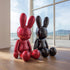 Two Avant-Garde Giant Seated Rabbit Fiberglass Sculpture in red and black color on a wooden floor with a large window view of a beach.