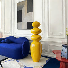 A Modern Glossy Yellow Stacked Pebble Fiberglass Sculpture in a 
Modern living room with a blue sofa, and abstract art on the wall.