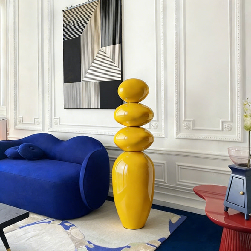 A Modern Glossy Yellow Stacked Pebble Fiberglass Sculpture in a 
Modern living room with a blue sofa, and abstract art on the wall.