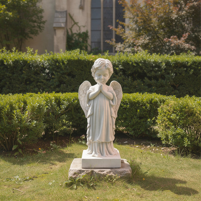 Gentle Praying Cherub Marble Sculpture in a garden setting with greenery and a building in the background