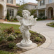 White marble cherub angel statue holding a lyre, displayed in a villa garden with lawn, flowers, and luxury home background.