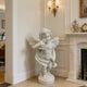 Natural white marble cherub angel statue placed indoors near a fireplace, adding a soft classical accent to elegant home decor.