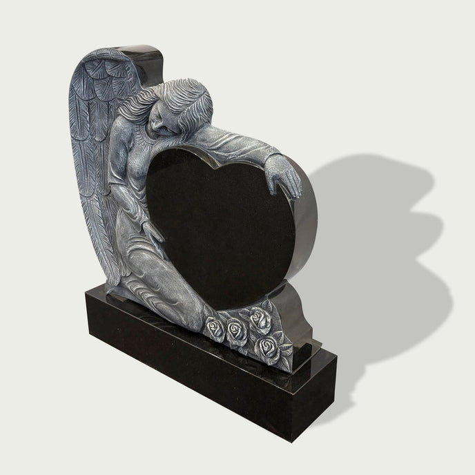 Side view of kneeling angel heart gravestone with black heart-shaped plaque, carved roses, feathered wings, and polished black stone base.