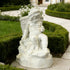 White marble child garden statue displayed near hedges and villa walkway, featuring a woven basket, small cup, and relaxed seated pose.