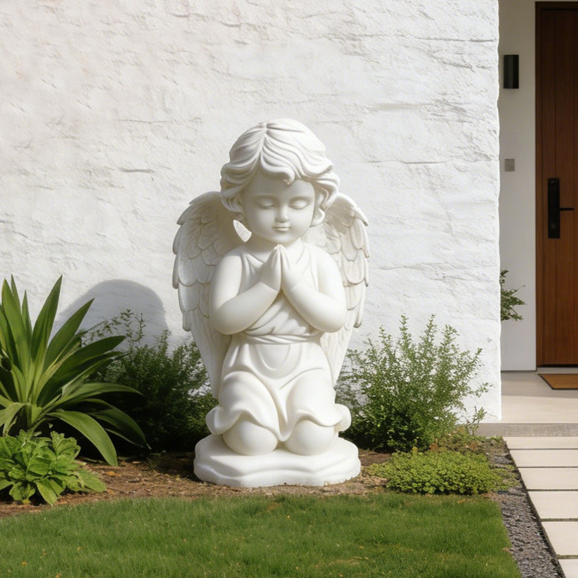 White marble kneeling angel statue placed near a modern garden wall, with folded hands, closed eyes, and calm devotional expression.