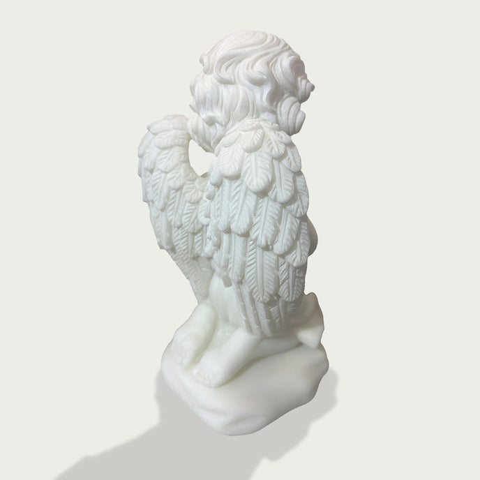 Side back view of white marble praying cherub angel sculpture showing layered wing carvings, curled hair, and kneeling position.