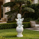 A white outdoor garden statue of a cherub seated on a round pedestal, placed on a green lawn in a landscaped courtyard with hedges, trees, patio furniture, and a building in the background.