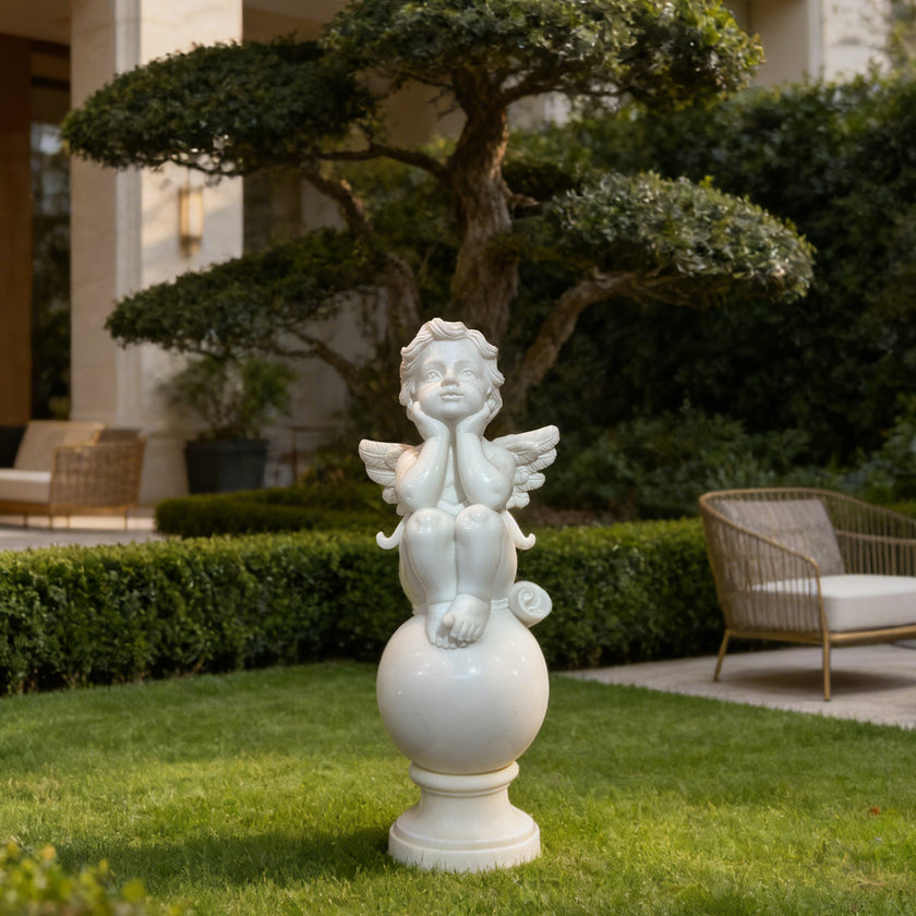 A white outdoor garden statue of a cherub seated on a round pedestal, placed on a green lawn in a landscaped courtyard with hedges, trees, patio furniture, and a building in the background.