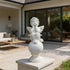 A white cherub statue seated on a round pedestal, placed on a stone planter in a modern patio setting with a house and outdoor furniture in the background.