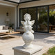 A white cherub statue seated on a round pedestal, placed on a stone planter in a modern patio setting with a house and outdoor furniture in the background.