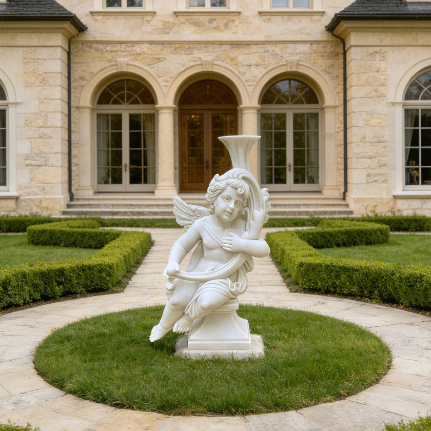 A white marble-style cherub garden statue holding a classical flute, set on a circular green lawn in front of a large stone mansion with arched windows.
