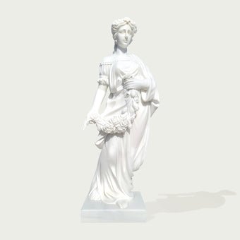 A white classical-style statue of a draped female figure standing on a rectangular base, shown against a light gray studio background with a soft shadow.