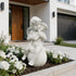 A white cherub garden statue sitting on a round base and holding a small object to its lips, placed in a landscaped bed with white flowers in front of a modern house entrance.