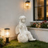 A white angel garden statue seated on a sunflower, placed on a lawn beside a house wall with a lit lantern and flower bed.