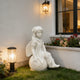 A white angel garden statue seated on a sunflower, placed on a lawn beside a house wall with a lit lantern and flower bed.