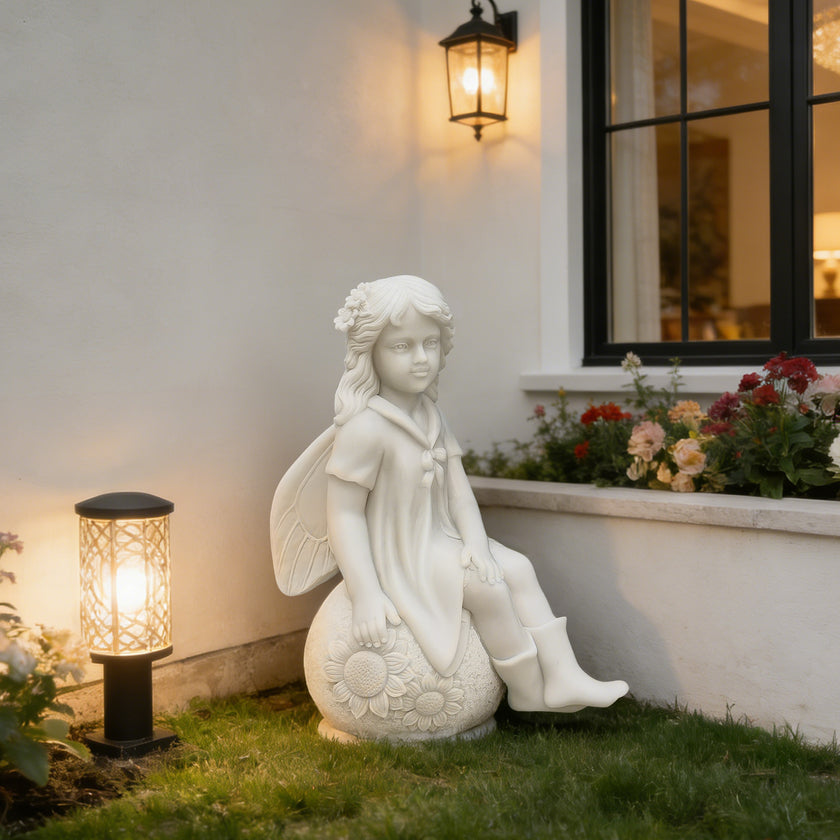 A white angel garden statue seated on a sunflower, placed on a lawn beside a house wall with a lit lantern and flower bed.