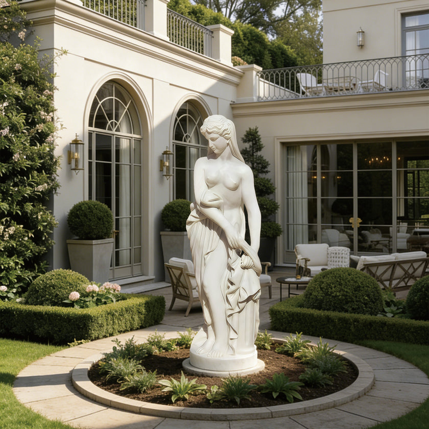 A white classical-style female statue stands in a landscaped garden courtyard in front of a large cream-colored house with arched windows and patio seating.