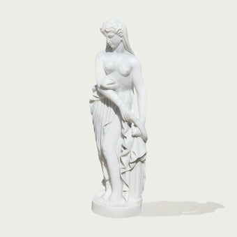 A white classical-style statue of a draped female figure standing on a round base, photographed against a plain light gray background with a soft shadow.