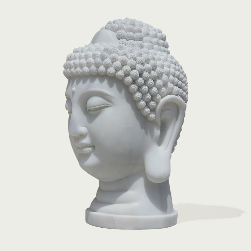 A white Buddha head bust sculpture with detailed textured hair, closed eyes, and a serene expression, shown on a light beige background.