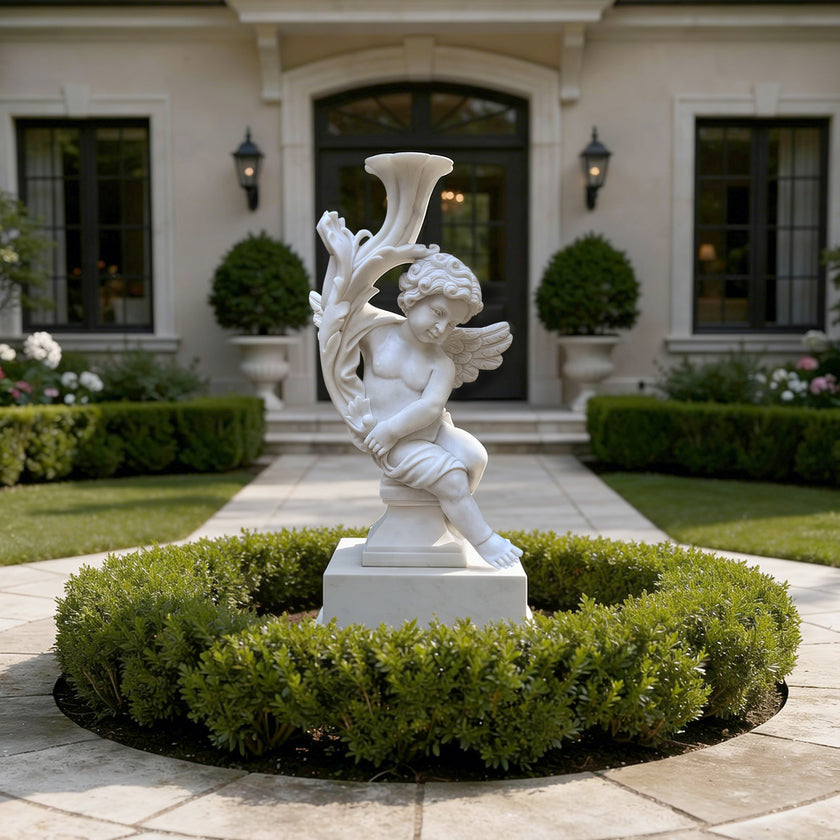 A white classical cherub garden statue seated on a base with a horn-like form, displayed in a landscaped courtyard with trimmed hedges and a stone walkway leading to a building entrance.