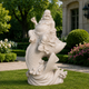 A white stone or resin statue of a smiling Buddha riding a fish, placed outdoors on a green lawn in front of a garden and building.