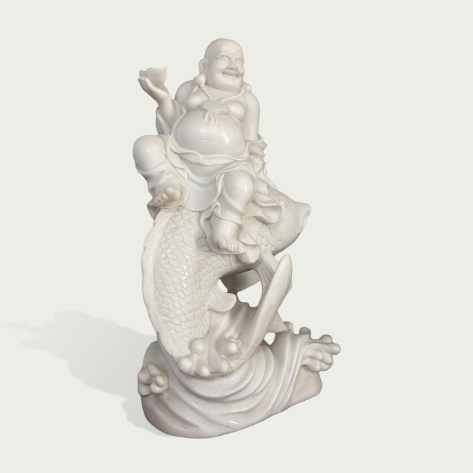 Side view of a white marble Laughing Buddha seated on a fish sculpture, with smooth handcrafted details and wave base design representing abundance and blessings.