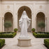 White marble religious woman statue displayed in a church courtyard, featuring flowing robes, open hands, detailed carving, and a peaceful sacred architectural setting.